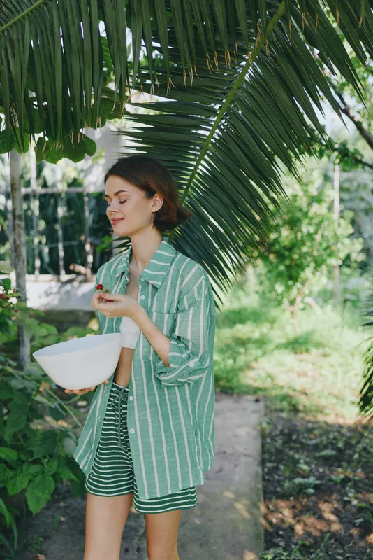 Young woman outdoors under tropical palm shade wearing a green striped shirt and shorts, holding a bowl, capturing casual summer style and relaxed mood.