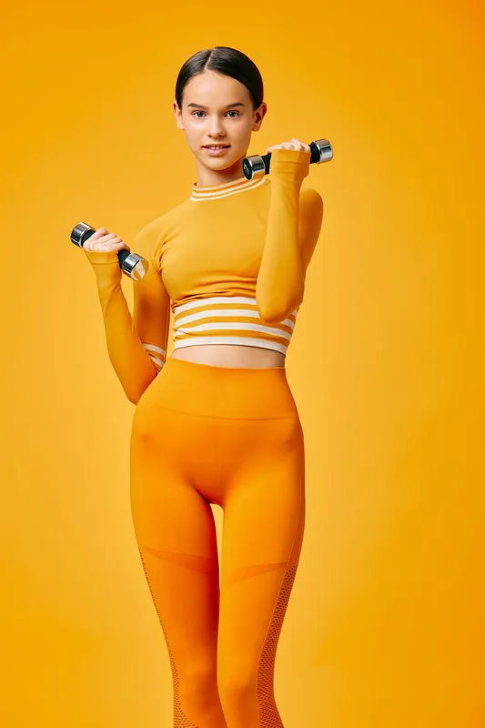 Female fitness model in bright orange activewear, holding dumbbells in a studio with a vivid orange backdrop, showcasing strength, balance and confident training.