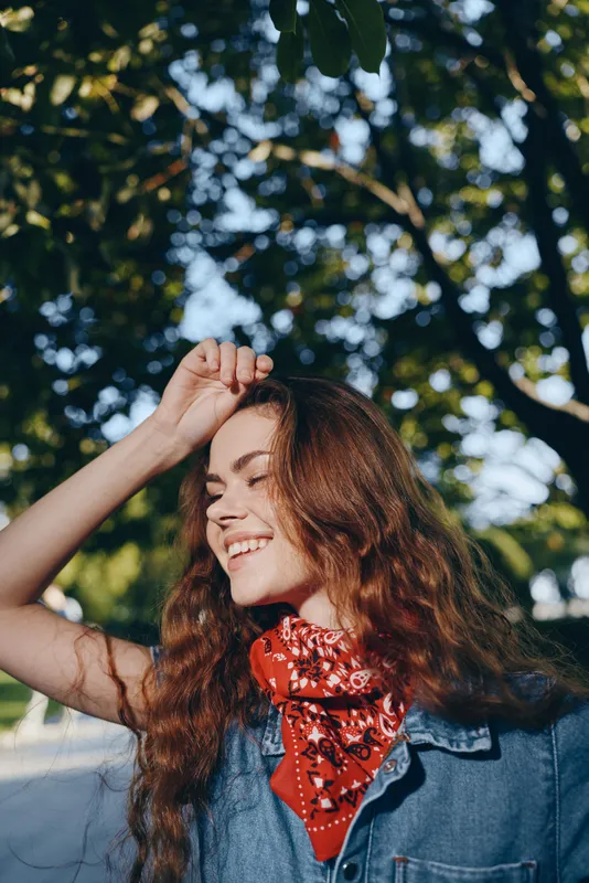 Lifestyle woman in modern boho-western denim street style smiling with red bandana and sunlit curly hair, outdoors with green foliage, vibrant film color tones.