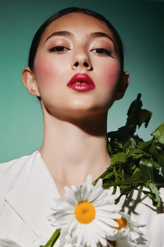 Young woman with glossy red lips and bold blush poses confidently holding green foliage with white flowers. Studio shot with artistic lighting emphasizing beauty and fashion concept.