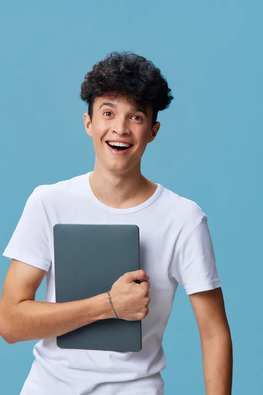 smiling young man holding laptop, wearing white t-shirt with curly hair against a blue background, casual and cheerful expression, modern technology concept