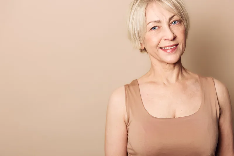 Smiling mature woman with short blonde hair wearing beige tank top stands against neutral background. Concept of confidence, aging, natural beauty, and positive lifestyle for older adults.