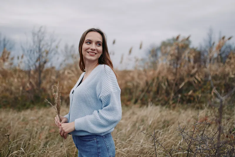 A candid portrait of a smiling young woman standing in tall grass, holding dried reeds and looking aside, conveying authenticity and a genuine connection to nature with relaxed posture and warm