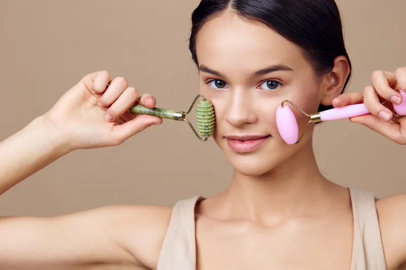 Young woman using facial rollers for skincare, showcasing a healthy beauty routine with green and pink rollers against a soft neutral background