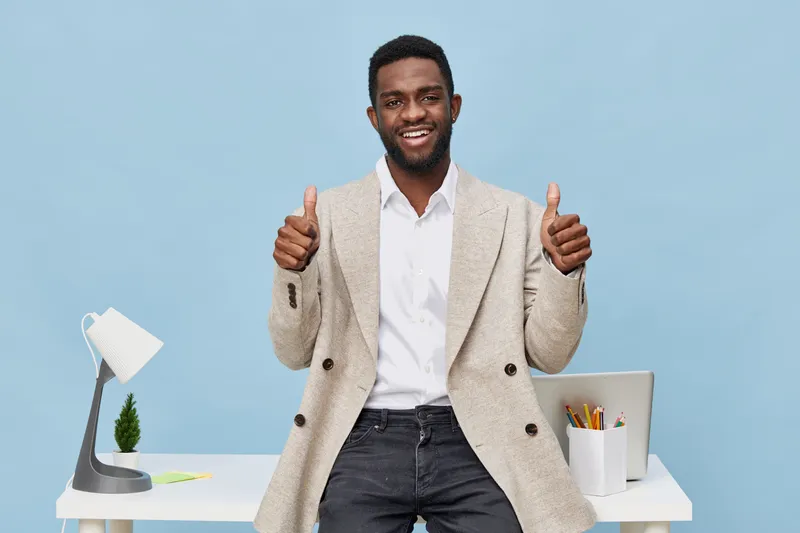Confident smiling man in casual blazer and white shirt giving thumbs up while standing in bright modern office with desk, lamp, and stationery on pastel blue background. Business success concept, prof