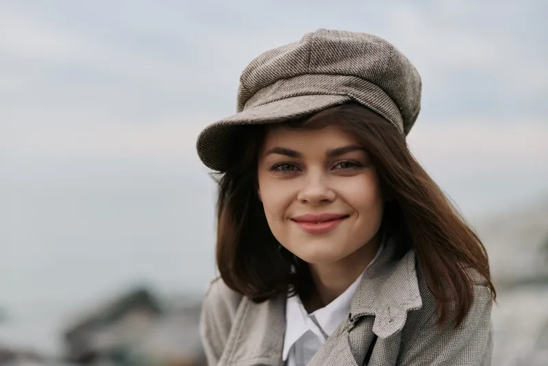 Young woman with brown hair wearing a grey hat and coat smiles softly outdoors. Blurred natural background creates a calm and relaxed mood. Concept of autumn fashion and lifestyle.