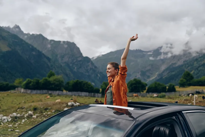 Outdoors woman stands through a car roof with one arm raised in celebration, amid dramatic mountains and an expansive sky, expressing travel adventure, freedom and a vivid landscape.