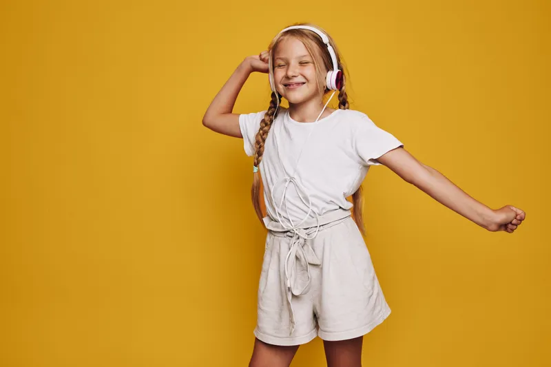 Happy girl with long braids listens to music with headphones and dances on yellow background. Smiling child in casual outfit enjoying sound and movement.