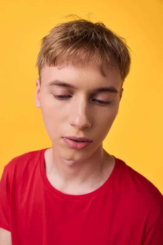 Close up of a young male in a red shirt, contemplative expression against a bright yellow backdrop, conveying introspection and focus for fashion or lifestyle mood