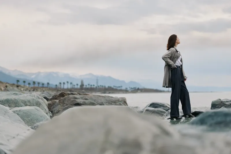 A woman stands on rocky shore looking into the distance with mountains and cloudy sky in the background. Concept of contemplation, nature, and outdoor exploration.