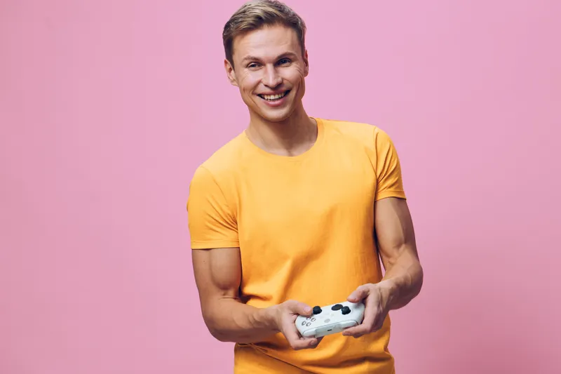 smiling young caucasian man hold game controller wear yellow t-shirt isolated on pink background casual lifestyle concept