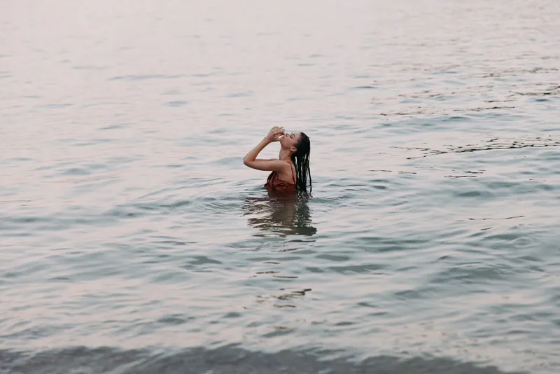 A person stands waist deep in calm water, tilting head back as hands frame the face, capturing a peaceful moment of solitude and refreshment.