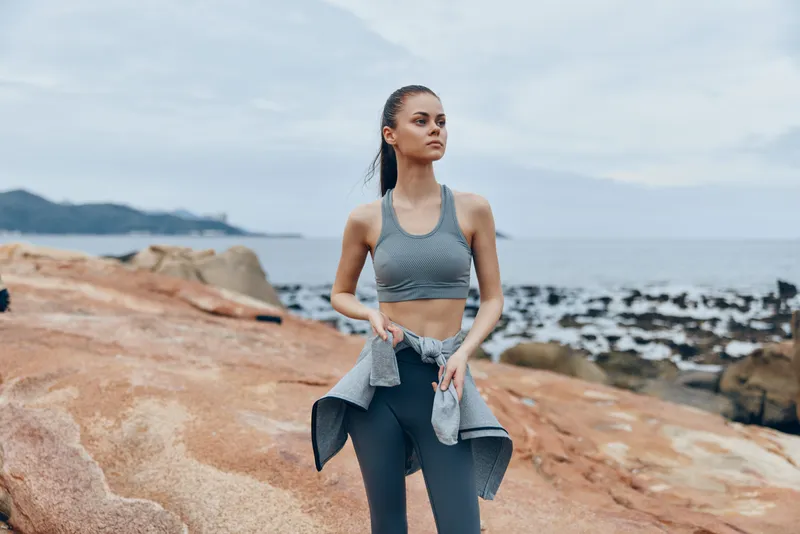 Young woman in athletic wear practicing mindfulness by the seaside, emphasizing health and wellness, dressed in a gray sports bra and leggings