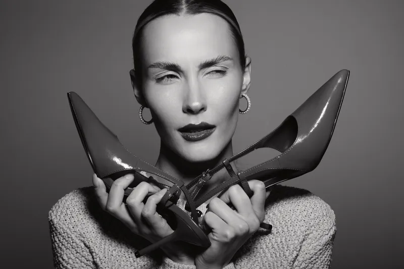 White woman with light skin holds two high heel shoes near face, posing in studio with serious expression. Fashion and style concept.