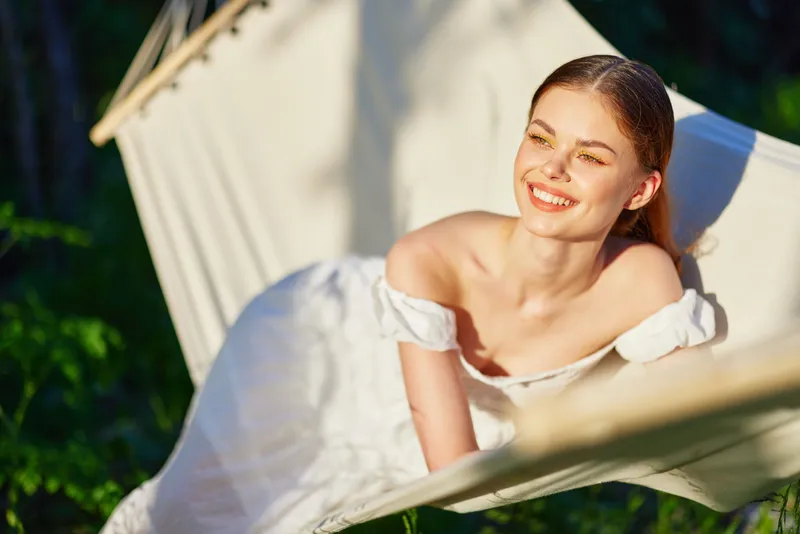 Beautiful young woman relaxing in a hammock outdoors, wearing a white dress, smiling happily in the sunshine, surrounded by lush green foliage