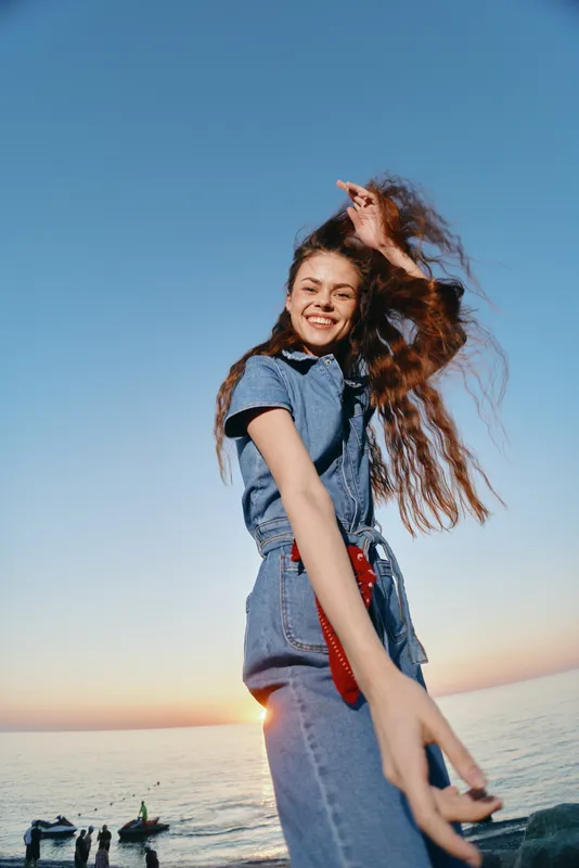 smiling woman in denim outfit standing by the ocean during sunset with long hair and joyful expression
