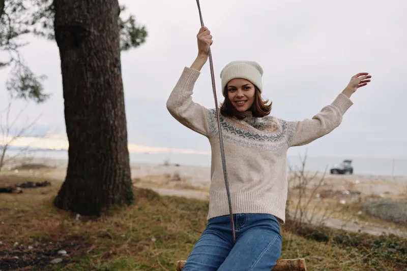 Woman on a swing outdoors, wearing a cozy sweater and beanie, smiling as she enjoys nature near the beach. Casual leisure scene with trees and open sky, capturing a playful mood.