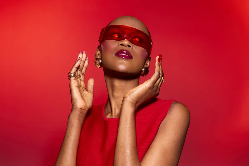 Bald black woman with dark skin wearing red sunglasses and sleeveless top poses against vibrant red background. Fashion portrait with confident expression and bold makeup for style concept.