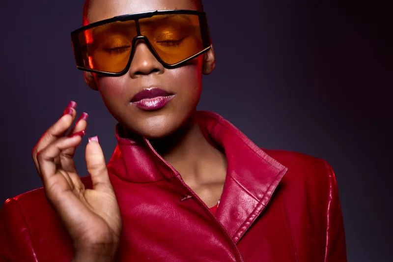 Close-up of an elegant black woman with dark skin wearing oversized orange sunglasses and a red leather jacket. Concept of fashion, style, confidence, and modern urban look.