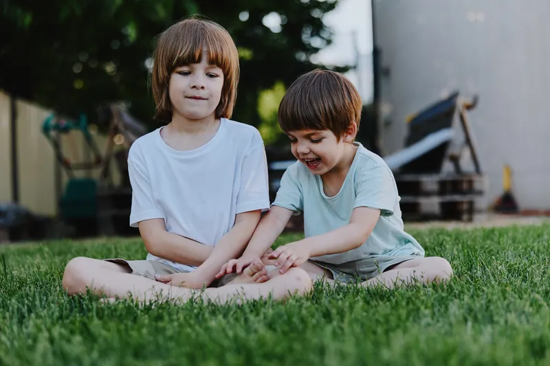 Two boys sitting on a grassy lawn, engaging in playful activity. One boy smiles while the other looks thoughtful, showcasing childhood innocence and joy.