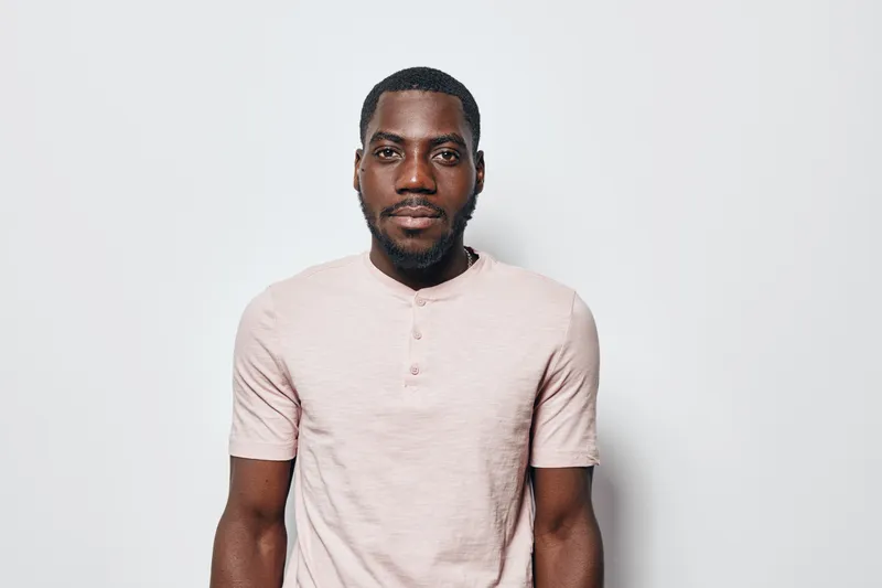African American man with dark skin and beard wearing light pink t shirt standing against white background looking at camera with neutral expression and confident posture.