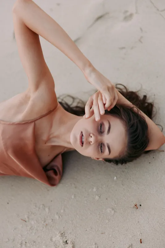 Portrait of a woman lying on warm sand with sunlit skin, soft fabric draped, and a relaxed pose; a minimal, elegant beach scene that conveys calm and natural beauty.