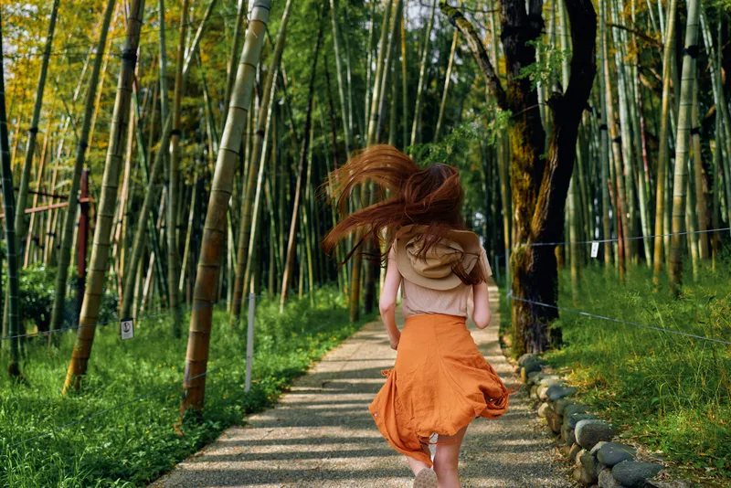 Woman running on a path through bamboo forest with backpack and orange skirt, rear view motion, sunlight filtering through tall stalks, outdoor trail adventure and carefree travel.