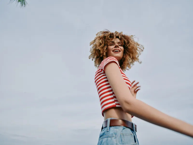 Young woman with curly hair wears a red and white striped crop top and denim jeans, smiling against a clear blue sky, capturing a carefree fashion moment in sunshine.