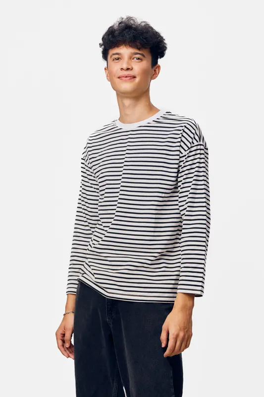 young man wearing striped long sleeve shirt standing relaxed against white background with casual black pants, natural expression, modern style portrait