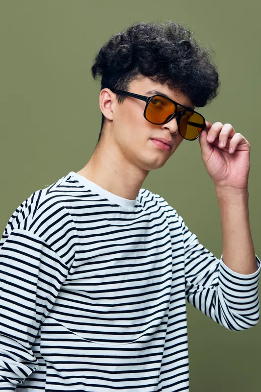 young man with sunglasses adjusting eyewear, curly hair, striped shirt, confident expression, studio portrait with green background, casual fashion style