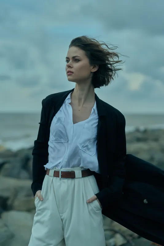 Windy coast scene with a woman in a blazer and white shirt, hands in pockets, looking into the distance, showcasing a modern business casual outfit on the windy shoreline
