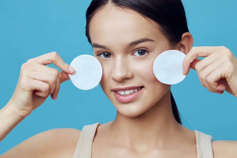 Young woman smiling while holding cotton pads, promoting skincare and beauty with a joyful expression against a light blue background