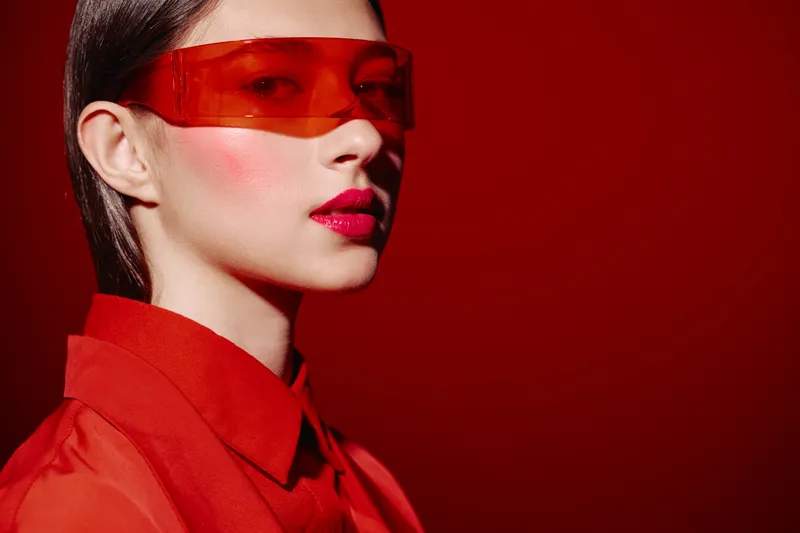 Young woman wearing red tinted glasses and red shirt poses with intense makeup and glossy lips on dark red background. Beauty fashion creative concept emphasizing bold look.