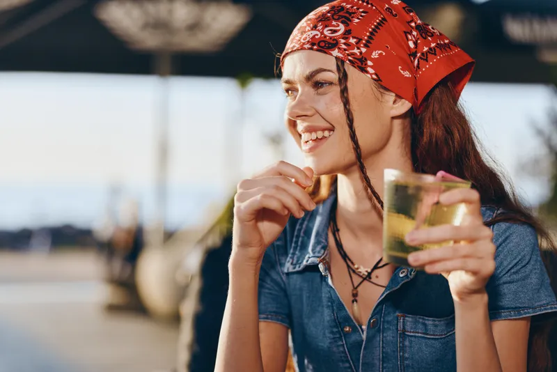 smiling woman with red bandana enjoying a drink outdoors on a sunny day with relaxed and happy mood near blurred background