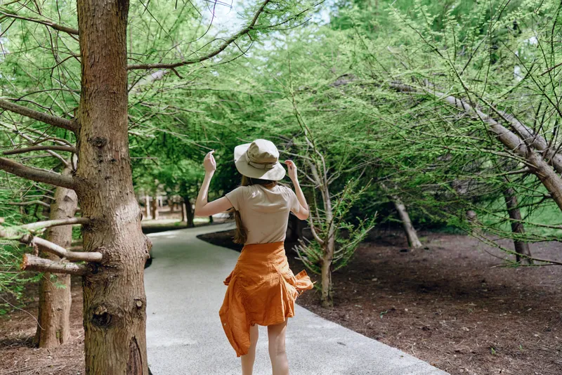 Woman walking in park under trees on a gravel pathway, wearing sunhat and orange skirt, casual summer leisure scene from rear with motion and carefree outdoor lifestyle.