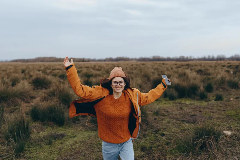 Smiling woman in an orange sweater stands in the grassy field, outdoor nature lifestyle with freedom and joy, arms raised for a celebratory dance, fresh air feeling in sunny afternoon