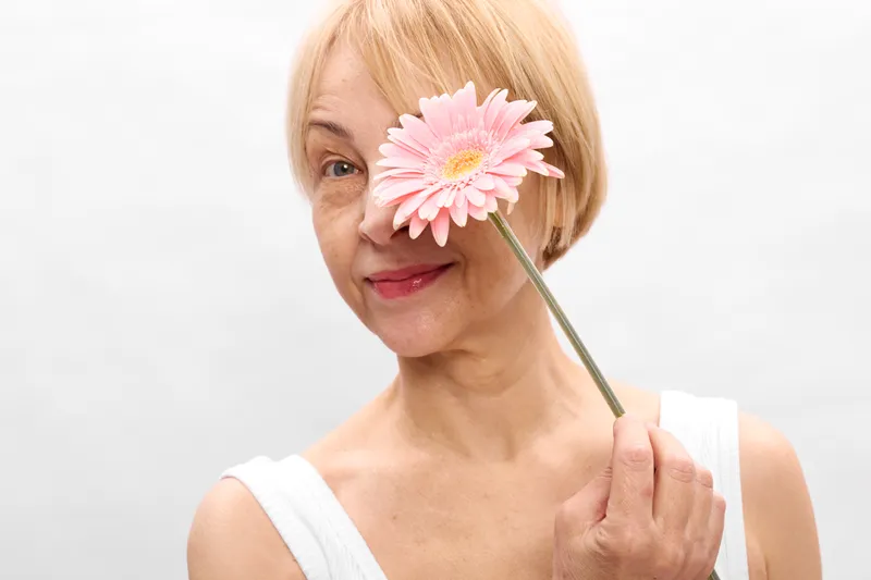 woman elderly portrait beauty flower skincare wellness cosmetic spring smile, a blonde elderly woman holds a pink daisy on a stick, soft studio light and calm facial expression.
