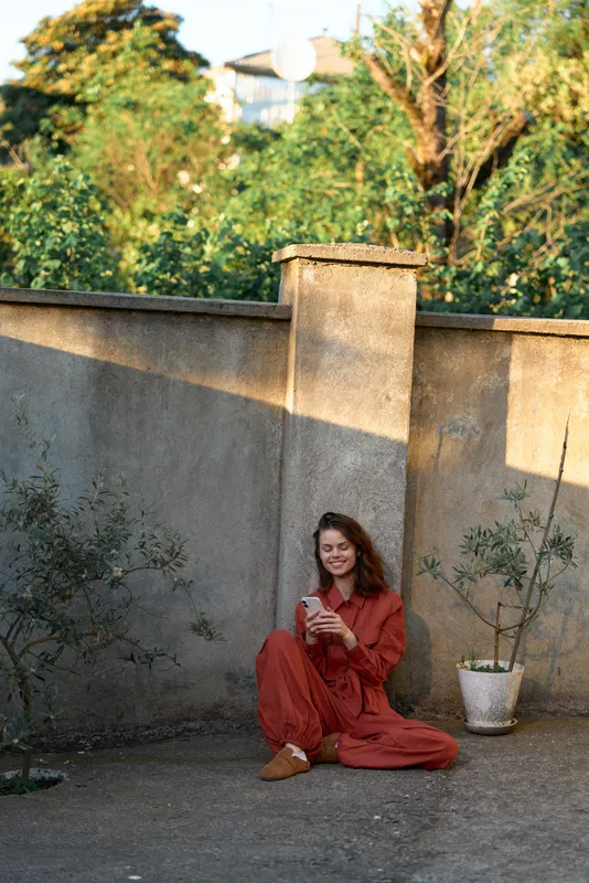 Young woman in a red dress sits on the ground outdoors beside a concrete wall, reading a book with a gentle smile and relaxed posture