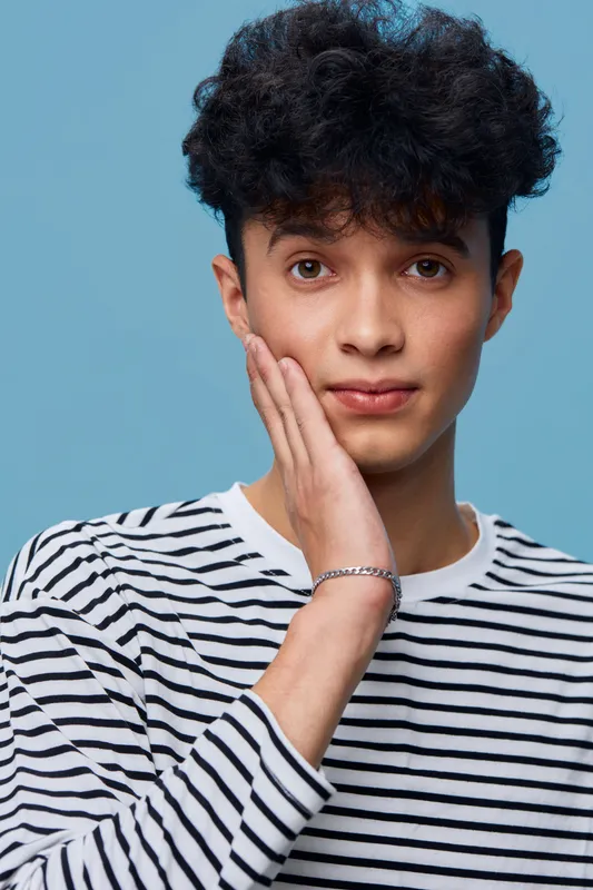 young man with curly hair touching face wearing striped shirt, thoughtful expression on blue background in casual style and soft lighting