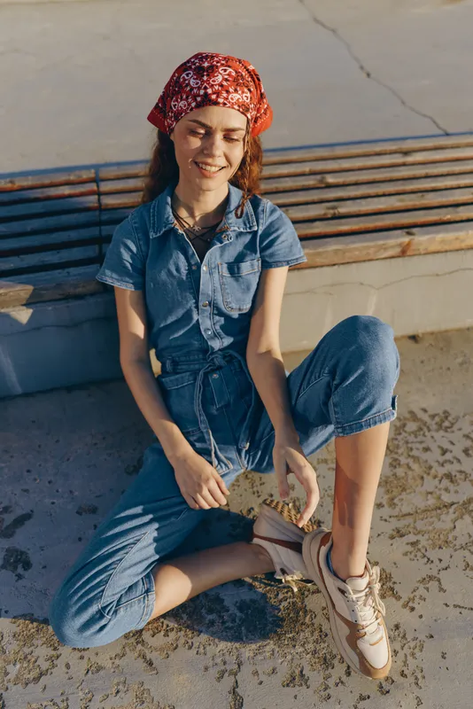 Lifestyle woman in modern boho-western denim street style wearing a red bandana and sneakers, smiling with relaxed posture in warm film color tones.