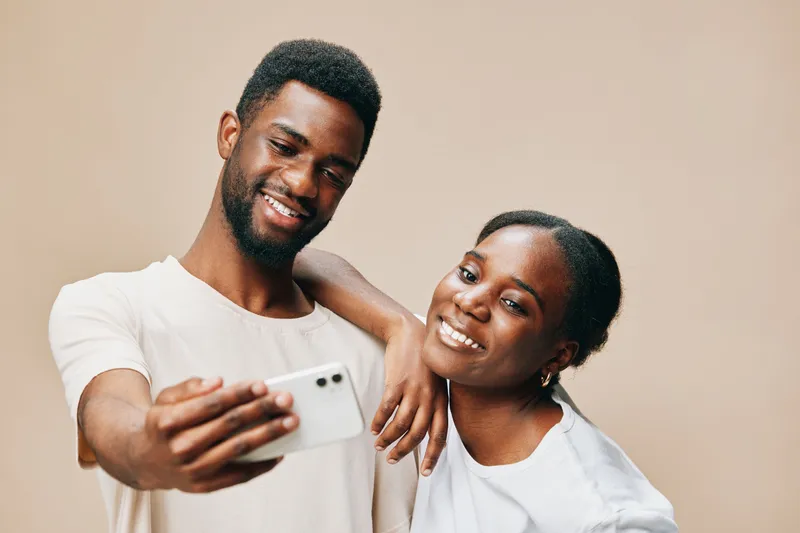 Smiling young African couple taking a selfie together against a neutral beige background, showcasing their happiness, love, and connection in a warm, intimate moment