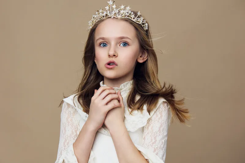 A surprised young girl in a white lace dress and crown, expressing wonder against a soft beige background, perfect for children s fashion and fairy tale concepts