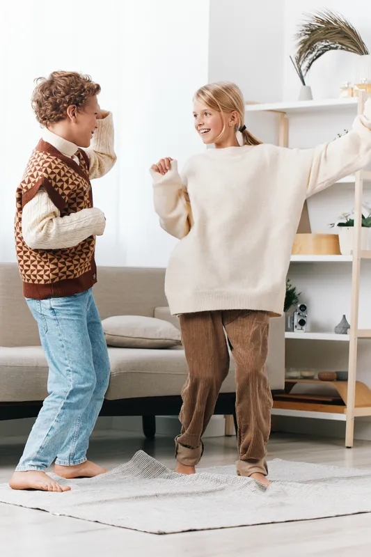 Two children dance happily indoors in a bright living room, wearing cozy sweaters and denim. Their playful energy and warm smiles create a cheerful, candid moment of everyday joy.