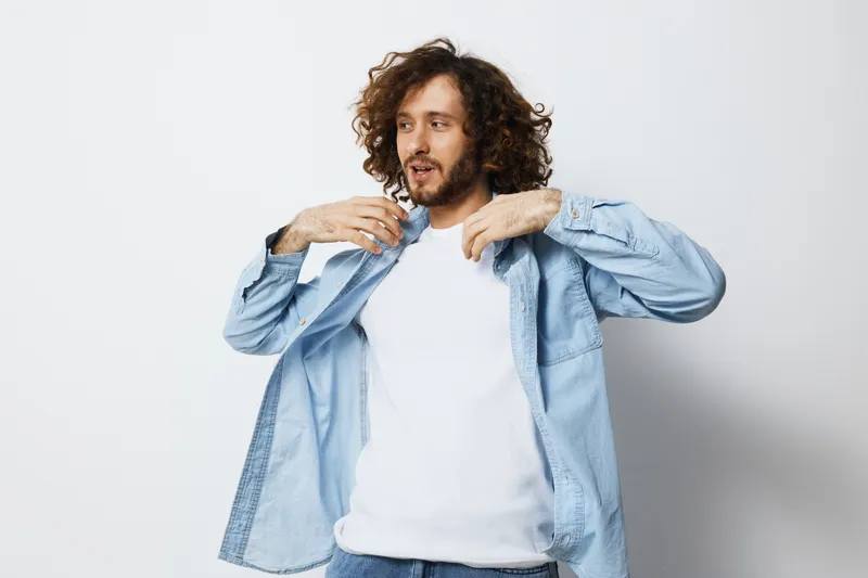 Young Caucasian man adjusting his denim shirt, expressing a casual and confident demeanor in a minimalistic white background Fashionable and modern look
