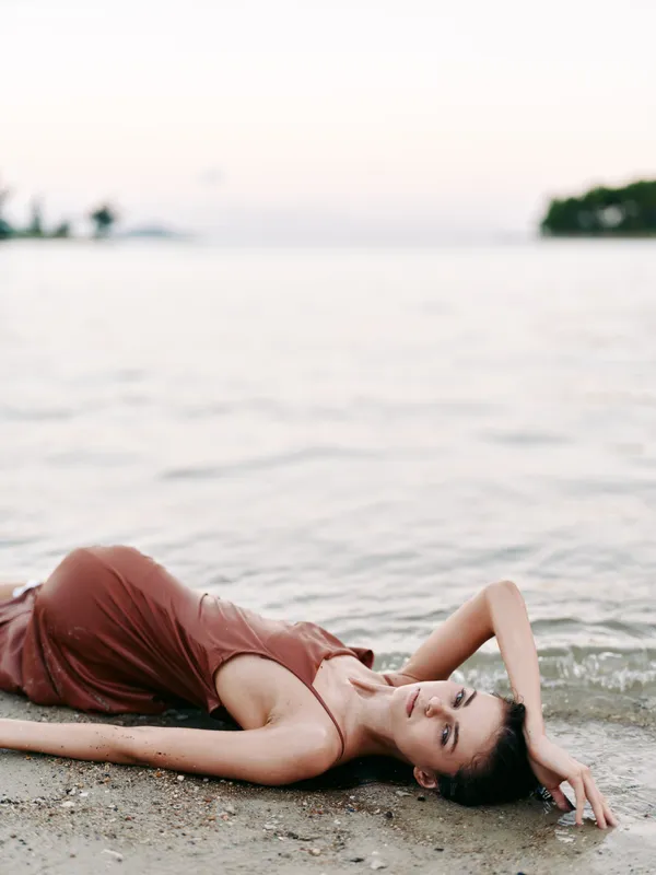 A relaxed woman lies on a sandy beach near gentle waves, wearing a soft red dress, eyes closed, creating a calm, intimate moment by the shoreline at sunset.