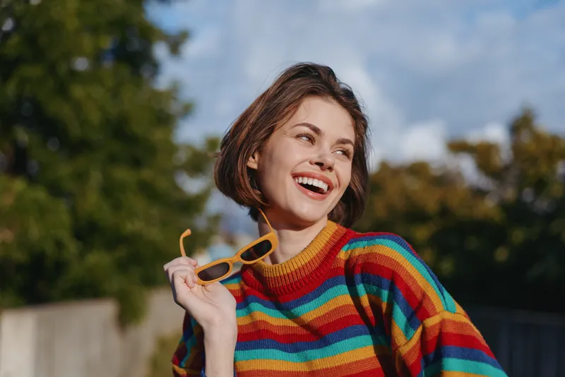 woman smile rainbow sweater sunglasses portrait outdoor enjoying sunlight, young woman laughing in colorful knit jumper with joyful expression and casual fashion vibe
