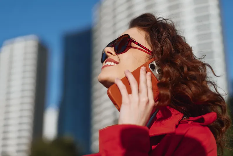 Young woman with curly hair wearing sunglasses and a red coat smiles while talking on a smartphone outdoors in an urban environment on a sunny day.