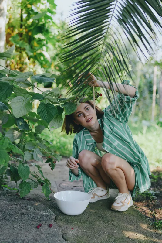 woman crouching under lush foliage, harvesting fruit with a white bowl nearby, in casual green striped shirt.