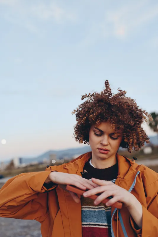 Woman with curly hair outdoors, wearing a brown jacket, holding a phone and texting at sunset, relaxed lifestyle, warm light, city background, soft wind