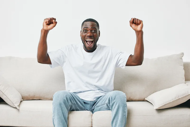 Happy Black man celebrating on a light colored couch, expressing joy and excitement, wearing a casual white t shirt, symbolizing victory and bliss